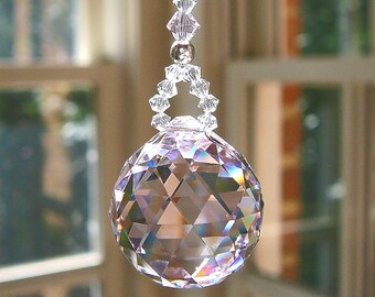 Op de afbeelding: Een heldere, veelzijdige kristallen bol die hangt aan een snoer van kleinere, heldere, geometrische kristallen kralen. De bol heeft een complex, driehoekig geslepen patroon dat het licht breekt en subtiele regenboogprisma's op het oppervlak creëert.