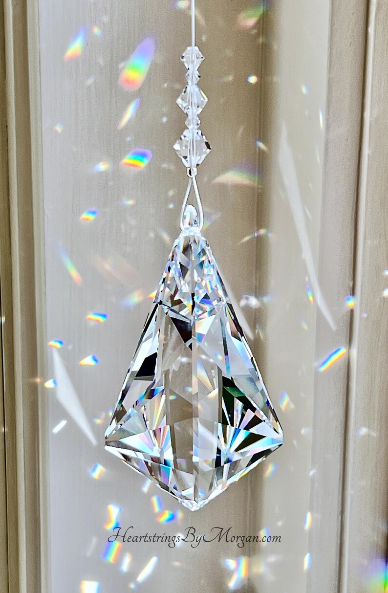 Puede incluir: Un prisma de cristal transparente y facetado suspendido de un hilo blanco. El prisma en forma de diamante tiene m&uacute;ltiples superficies reflectantes, creando patrones de luz de arco&iacute;ris en la pared. El texto "Heartstrings By Morgan.com" es visible.