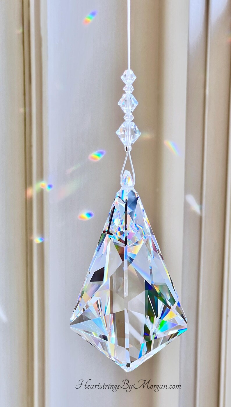 Puede incluir: Un atrapasoles de prisma de cristal transparente y facetado con un dise&ntilde;o geom&eacute;trico, suspendido por un fino hilo blanco. El prisma es triangular con una base en forma de diamante. La imagen muestra refracciones de luz arco&iacute;ris. Texto: Heartstrings By Morgan.