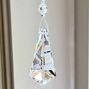 May include: A clear crystal teardrop prism hanging from a white string with clear crystal beads. The prism has a faceted design and catches the light, creating rainbows. The text "Heartstrings By Morgan.com" is visible at the bottom of the image.