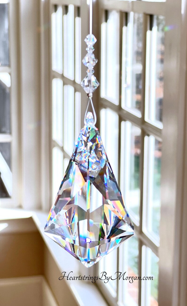 Puede incluir: Un atrapasol de prisma de cristal transparente y facetado, suspendido por un hilo fino. El prisma tiene forma de diamante con m&uacute;ltiples facetas, que reflejan un arco&iacute;ris de colores. El texto "Heartstrings By Morgan.com" es visible.