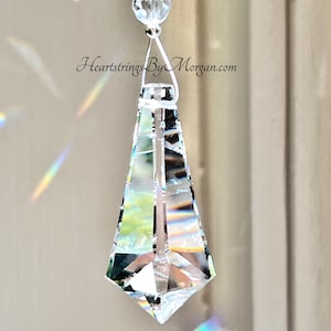 May include: A clear crystal prism hanging from a silver chain with a silver bead at the top. The prism is shaped like a teardrop and has a pointed tip. The prism is casting rainbows on the wall behind it. Heartstrings By Morgan.com is printed on the wall.