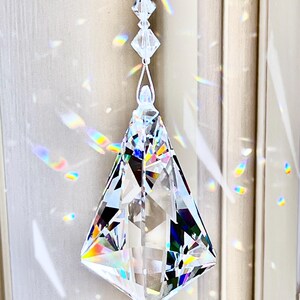 Puede incluir: Un atrapasoles de prisma de cristal transparente y facetado cuelga de un hilo blanco. El prisma tiene forma de diamante y refleja un arco&iacute;ris de colores. El texto "Heartstrings By Morgan.com" es visible.