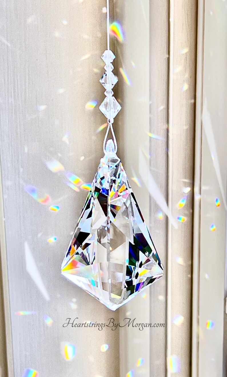 Puede incluir: Un atrapasoles de prisma de cristal transparente y facetado cuelga de un hilo blanco. El prisma tiene forma de diamante y refleja un arco&iacute;ris de colores. El texto "Heartstrings By Morgan.com" es visible.