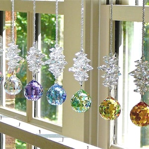 May include: A collection of hanging crystal suncatchers in various colors, including black, teal, purple, and yellow. Each suncatcher has a faceted crystal ball suspended from a decorative silver chain, reflecting light.