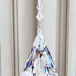 Puede incluir: Un prisma de cristal transparente con forma geom&eacute;trica cuelga de un fino hilo blanco. El prisma est&aacute; facetado, reflejando la luz y creando un efecto arco&iacute;ris. El texto "Heartstrings By Morgan.com" est&aacute; en la parte inferior.