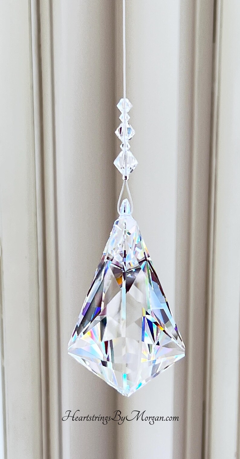 Puede incluir: Un prisma de cristal transparente con forma geom&eacute;trica cuelga de un fino hilo blanco. El prisma est&aacute; facetado, reflejando la luz y creando un efecto arco&iacute;ris. El texto "Heartstrings By Morgan.com" est&aacute; en la parte inferior.
