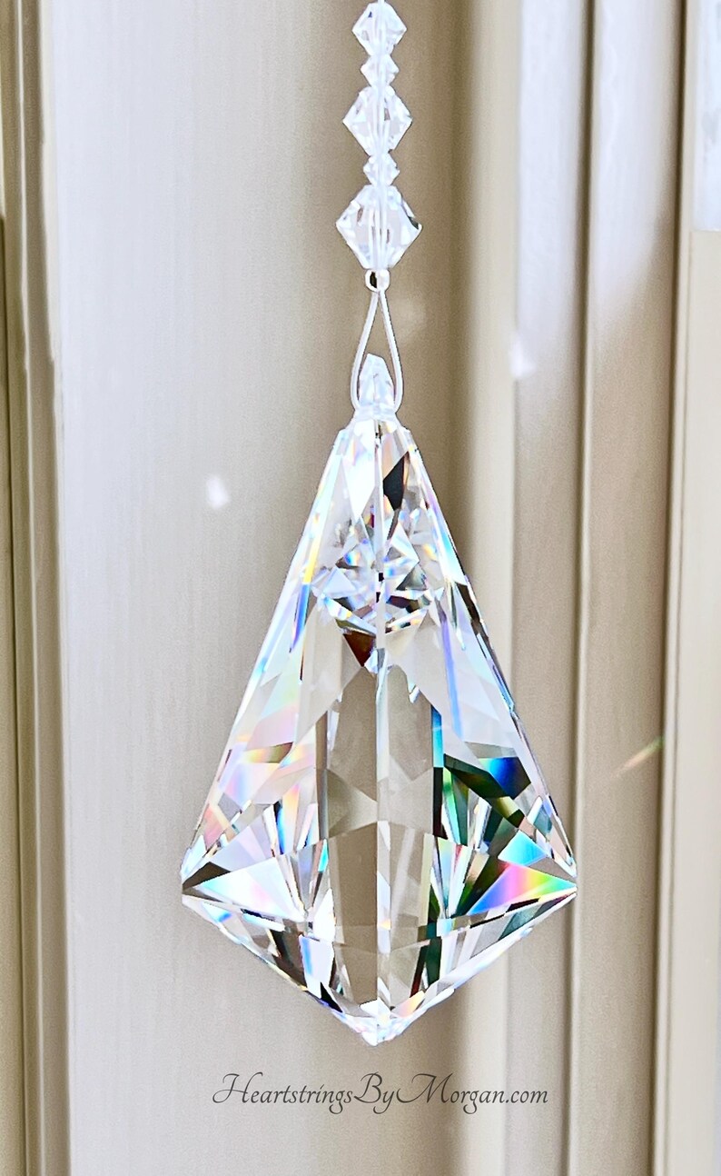 Puede incluir: Un colgante de prisma de cristal transparente y facetado con forma geom&eacute;trica, suspendido por una cadena de cuentas transparentes. El prisma refleja un arco&iacute;ris de colores. La direcci&oacute;n del sitio web Heartstrings By Morgan.com es visible.
