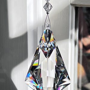 Puede incluir: Un atrapasoles de prisma de cristal transparente y facetado, suspendido por una cadena de cristales m&aacute;s peque&ntilde;os. El prisma tiene forma de diamante y refleja un arco&iacute;ris de colores. El texto "Heartstrings By Morgan.com" es visible.