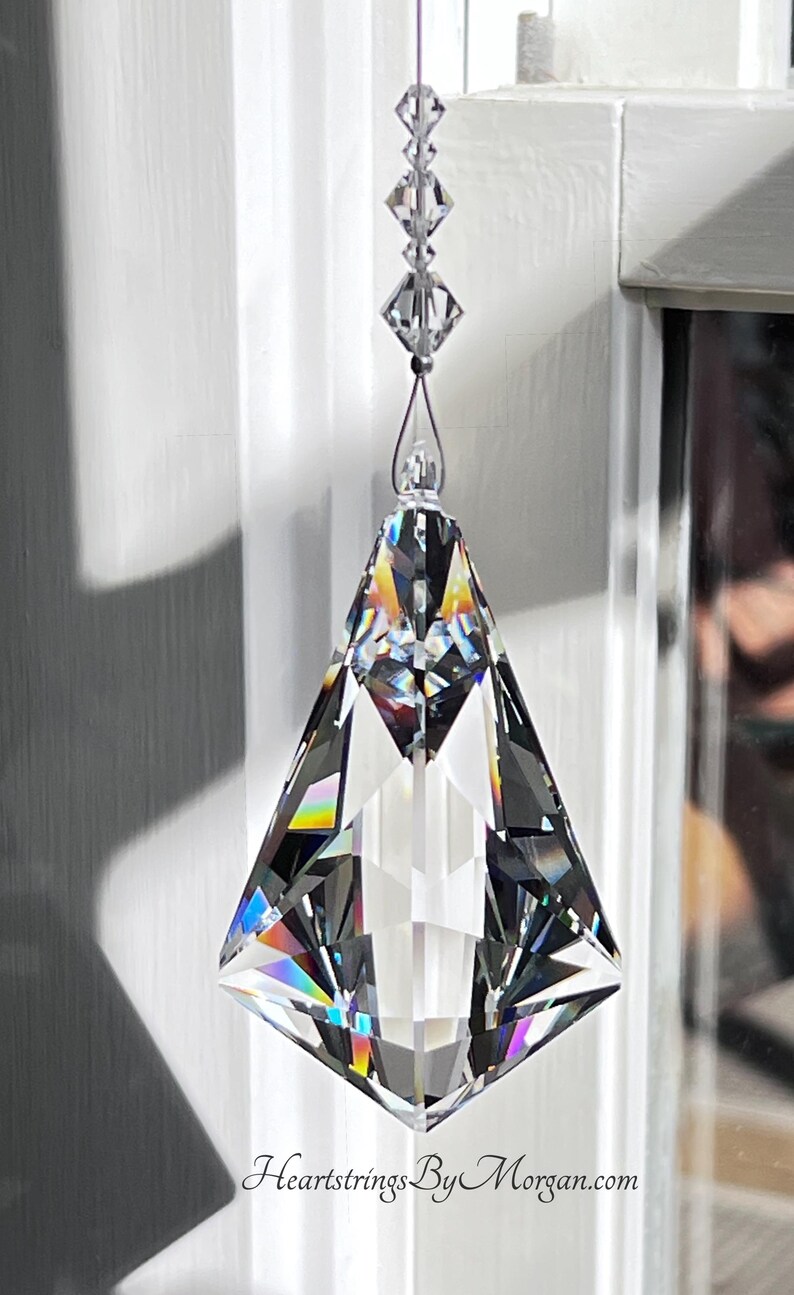 Puede incluir: Un atrapasoles de prisma de cristal transparente y facetado, suspendido por una cadena de cristales m&aacute;s peque&ntilde;os. El prisma tiene forma de diamante y refleja un arco&iacute;ris de colores. El texto "Heartstrings By Morgan.com" es visible.