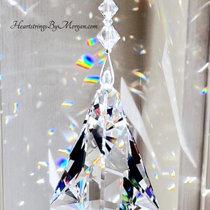 Puede incluir: Un atrapasoles de prisma de cristal transparente y facetado, suspendido por un hilo fino. El prisma tiene forma de diamante con m&uacute;ltiples facetas, que reflejan arco&iacute;ris de luz. El texto "Heartstrings By Morgan.com" es visible.
