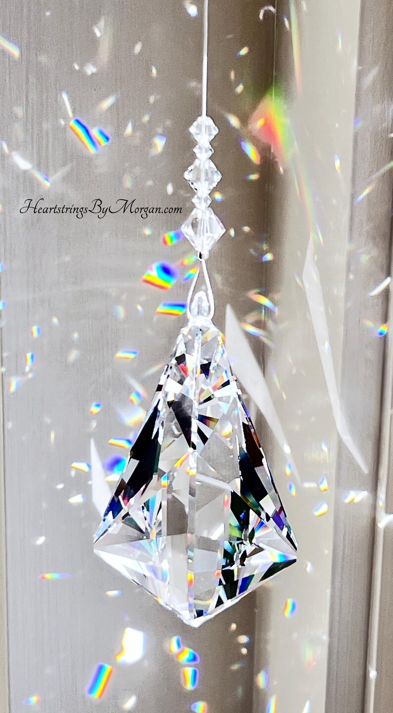 Puede incluir: Un atrapasoles de prisma de cristal transparente y facetado, suspendido por un hilo fino. El prisma tiene forma de diamante con m&uacute;ltiples facetas, que reflejan arco&iacute;ris de luz. El texto "Heartstrings By Morgan.com" es visible.
