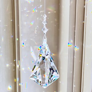 Puede incluir: Un atrapasol de prisma de cristal transparente y facetado suspendido por un hilo fino. El prisma tiene forma de diamante y refleja arco&iacute;ris de luz. La direcci&oacute;n del sitio web HeartstringsByMorgan.com es visible en la parte inferior.