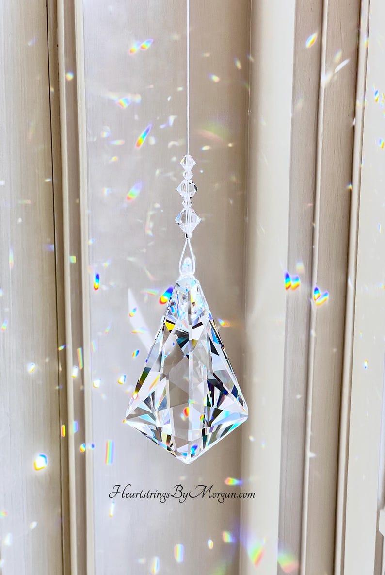 Puede incluir: Un atrapasol de prisma de cristal transparente y facetado suspendido por un hilo fino. El prisma tiene forma de diamante y refleja arco&iacute;ris de luz. La direcci&oacute;n del sitio web HeartstringsByMorgan.com es visible en la parte inferior.