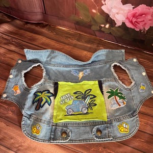 May include: A denim vest with various patches, including palm trees, bees, and a lightning bolt. A green pocket features a blue car and the words "GONE SURFIN". The vest has a button closure and is designed for a child.