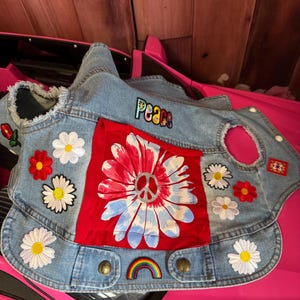 May include: A denim dog vest with a red and white daisy design and the word "Peace" embroidered on the back. The vest is decorated with daisy and rainbow patches, and has brass-colored buttons.