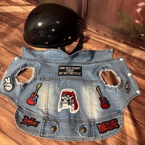 May include: A black motorcycle helmet rests above a light blue denim vest with patches. The vest has patches that say "Bon Jovi", "The Cure", and "Some Need Therapy I Have My Motorcycle". Patches include guitars, a skull, and peace signs.