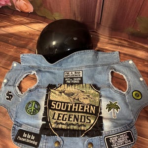 May include: A denim dog vest with patches, including a dollar sign, peace sign, and palm tree. The vest features the text "SOUTHERN LEGENDS" and other phrases. A black helmet rests behind the vest.