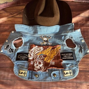 May include: A brown cowboy hat rests above a light blue denim vest adorned with patches. The vest features a skull and crossbones, a winged skull graphic, and the text "I'm Your Huckleberry". Patches with the word "Love" are also visible.