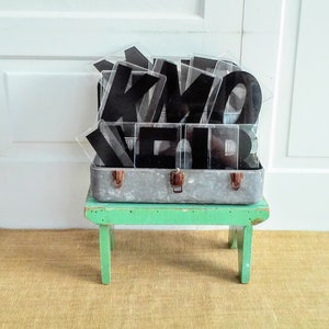 May include: A vintage metal suitcase filled with black plastic letters, sitting on a green wooden stool. The suitcase and letters are on a brown woven rug.