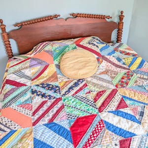 May include: A wooden headboard with turned posts and a decorative top rail sits behind a bed covered in a multicolored patchwork quilt featuring various fabric patterns and a single round tan pillow placed in the center of the bed.