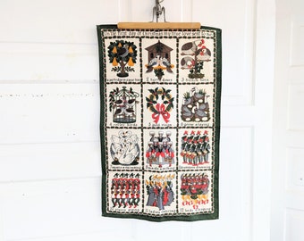 May include: A green fabric wall hanging with a white background and colorful illustrations of the 12 Days of Christmas. The text reads "On the first day of Christmas my true love sent to me..." and lists each day's gift.