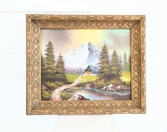 Puede incluir: Una pintura enmarcada que representa una escena de montaña nevada con una pequeña cabaña en primer plano. La pintura está en un marco dorado con detalles ornamentados.