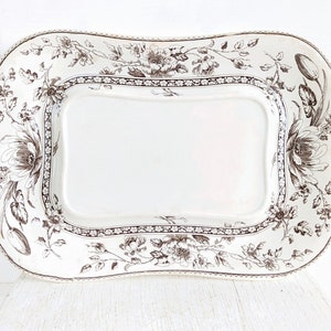 May include: A rectangular ceramic serving platter with rounded corners and a scalloped edge. The platter features a white base with a dark brown transferware floral pattern, including large blossoms, leaves, and a decorative border around the center.