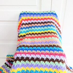 Op de afbeelding: Een kleurrijk gestreept gehaakt deken met verschillende kleuren, waaronder geel, blauw, rood, groen en paars.