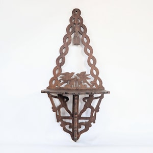 Op de afbeelding: Een donkerbruine houten wandplank met een ingewikkeld opengewerkt achterpaneel met een uitgesneden vogelmotief. De plank wordt ondersteund door een decoratieve beugel met twee figuren in profiel en heeft een hoog, gevlochten frame.