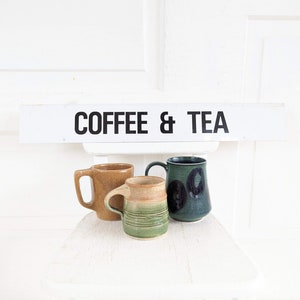 Puede incluir: Tres tazas de cer&aacute;mica, una marr&oacute;n, una verde con manchas negras y una verde con rayas marrones, sentadas en una silla blanca con un letrero arriba que dice "COFFEE & TEA".