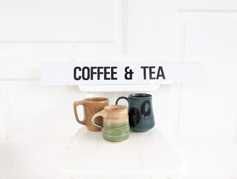 Puede incluir: Tres tazas de cer&aacute;mica, una marr&oacute;n, una verde con manchas negras y una verde con rayas marrones, sentadas en una silla blanca con un letrero arriba que dice "COFFEE & TEA".