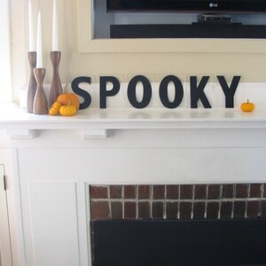 May include: A white fireplace mantel with a black brick surround and a black fireplace insert. The mantel is decorated with three wooden candle holders with white candles, three small orange pumpkins, and black wooden letters spelling "SPOOKY".