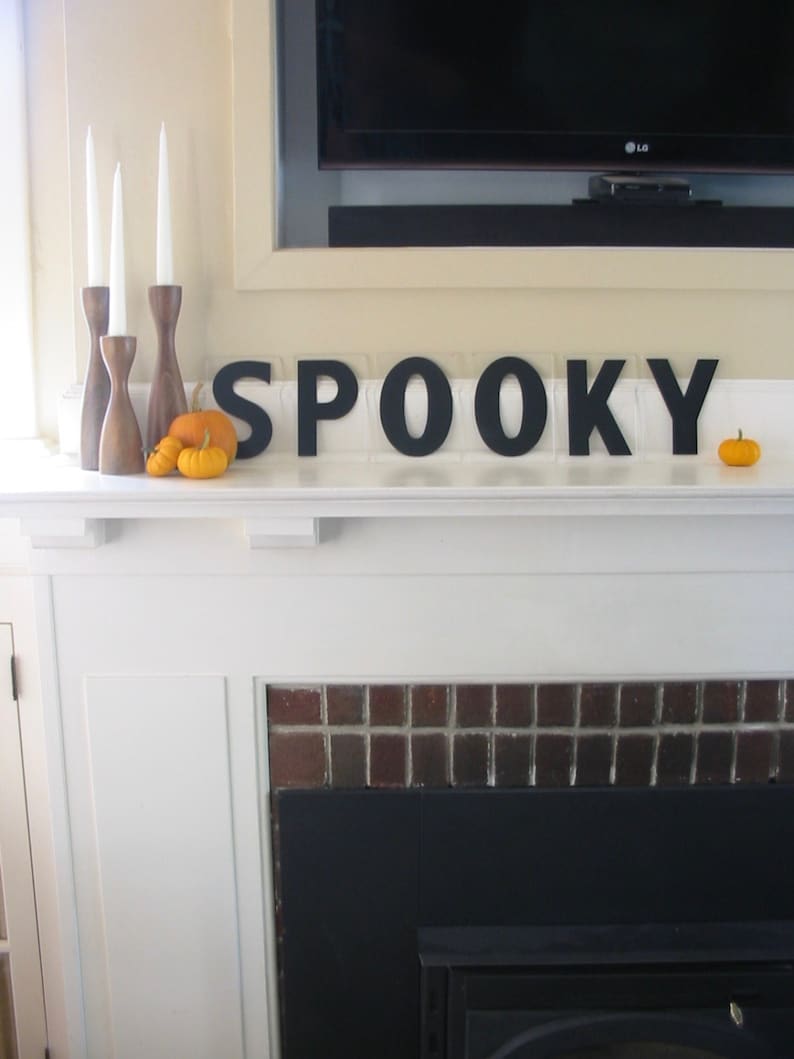 May include: A white fireplace mantel with a black brick surround and a black fireplace insert. The mantel is decorated with three wooden candle holders with white candles, three small orange pumpkins, and black wooden letters spelling "SPOOKY".