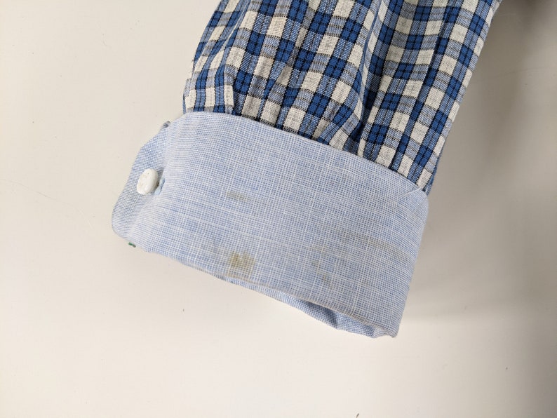 Op de afbeelding: Een blauw-wit geruit overhemd met een lichtblauwe manchet. De manchet heeft een witte knoop.