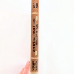 K&ouml;nnte beinhalten: Ein Vintage-Lehrbuch mit dem Titel "English Activities" f&uuml;r die achte Klasse. Das Buch hat einen braunen Einband mit wei&szlig;em Text und einen beigefarbenen Buchr&uuml;cken. Der Verlag ist die American Book Company. Das Buch wird von einer Hand gehalten.
