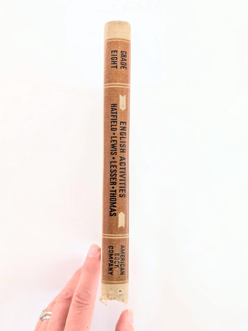 K&ouml;nnte beinhalten: Ein Vintage-Lehrbuch mit dem Titel "English Activities" f&uuml;r die achte Klasse. Das Buch hat einen braunen Einband mit wei&szlig;em Text und einen beigefarbenen Buchr&uuml;cken. Der Verlag ist die American Book Company. Das Buch wird von einer Hand gehalten.