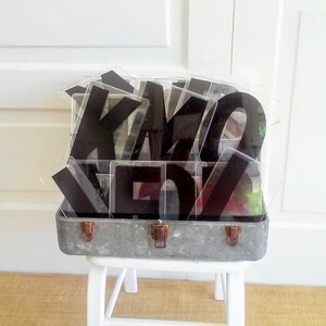 May include: A vintage metal suitcase filled with black acrylic letters. The letters are in a clear plastic container and spell out the word "KIMO"