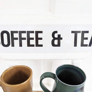 Puede incluir: Un letrero blanco con texto negro que dice "COFFEE & TEA". Dos tazas est&aacute;n en primer plano, una es marr&oacute;n y la otra es verde.