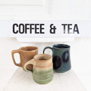 Puede incluir: Tres tazas de cer&aacute;mica, una marr&oacute;n, una verde con c&iacute;rculos negros y una verde y marr&oacute;n con un dise&ntilde;o a rayas. Las tazas est&aacute;n sobre una superficie blanca con un letrero encima que dice "COFFEE & TEA".