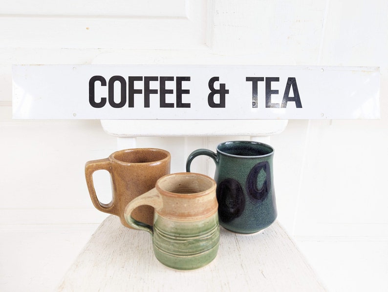 Puede incluir: Tres tazas de cer&aacute;mica, una marr&oacute;n, una verde con c&iacute;rculos negros y una verde y marr&oacute;n con un dise&ntilde;o a rayas. Las tazas est&aacute;n sobre una superficie blanca con un letrero encima que dice "COFFEE & TEA".