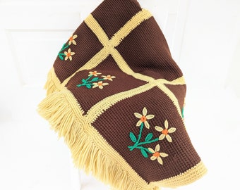Puede incluir: Una manta de crochet marrón con flecos amarillos y flores amarillas bordadas. La manta tiene un patrón de cuadros con cuadrados amarillos.