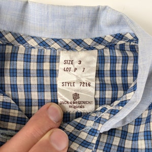 Op de afbeelding: Een blauw-wit geruit overhemd met een label met de tekst "SIZE 3 LOT P 7 STYLE 7216 JACK BORGENSICHT originals".