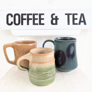Puede incluir: Tres tazas de cer&aacute;mica, una marr&oacute;n, una verde con manchas negras y una verde y marr&oacute;n con un dise&ntilde;o a rayas. Las tazas est&aacute;n sobre una superficie blanca con un cartel que dice "COFFEE & TEA".