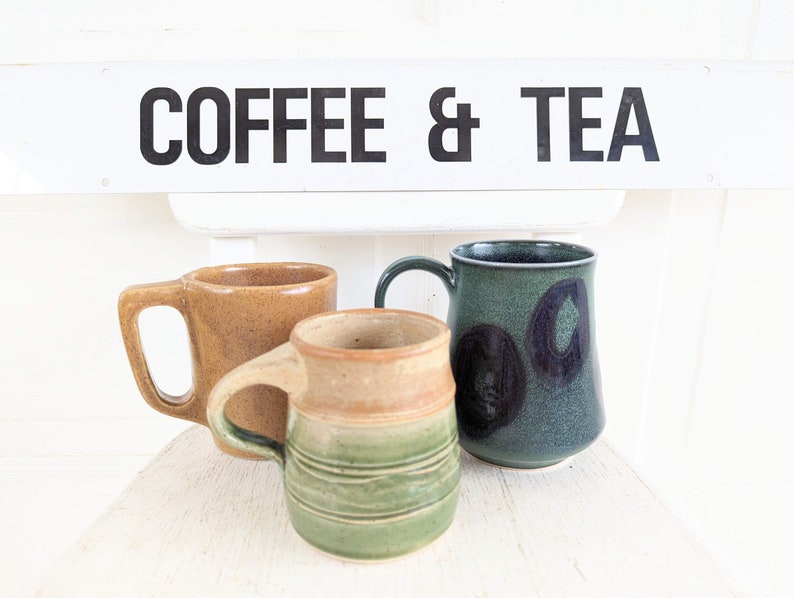 Puede incluir: Tres tazas de cer&aacute;mica, una marr&oacute;n, una verde con manchas negras y una verde y marr&oacute;n con un dise&ntilde;o a rayas. Las tazas est&aacute;n sobre una superficie blanca con un cartel que dice "COFFEE & TEA".