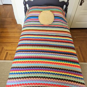 Op de afbeelding: Een kleurrijk gestreept gehaakt deken met een bruin rond kussen erop, liggend op een donker houten bedframe.