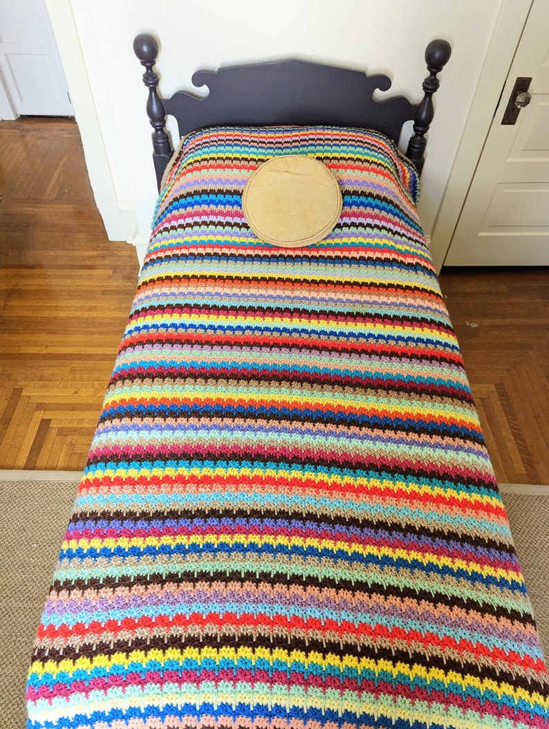 Op de afbeelding: Een kleurrijk gestreept gehaakt deken met een bruin rond kussen erop, liggend op een donker houten bedframe.