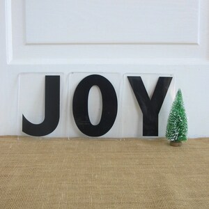 May include: Three black letters spelling "JOY" on a white background. The letters are made of a thick, black material and are set on a clear acrylic base. A small, green artificial Christmas tree is in the foreground.