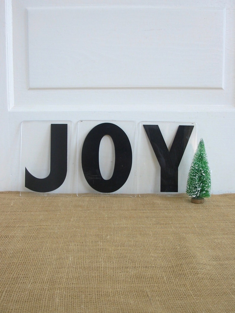 May include: Three black letters spelling "JOY" on a white background. The letters are made of a thick, black material and are set on a clear acrylic base. A small, green artificial Christmas tree is in the foreground.