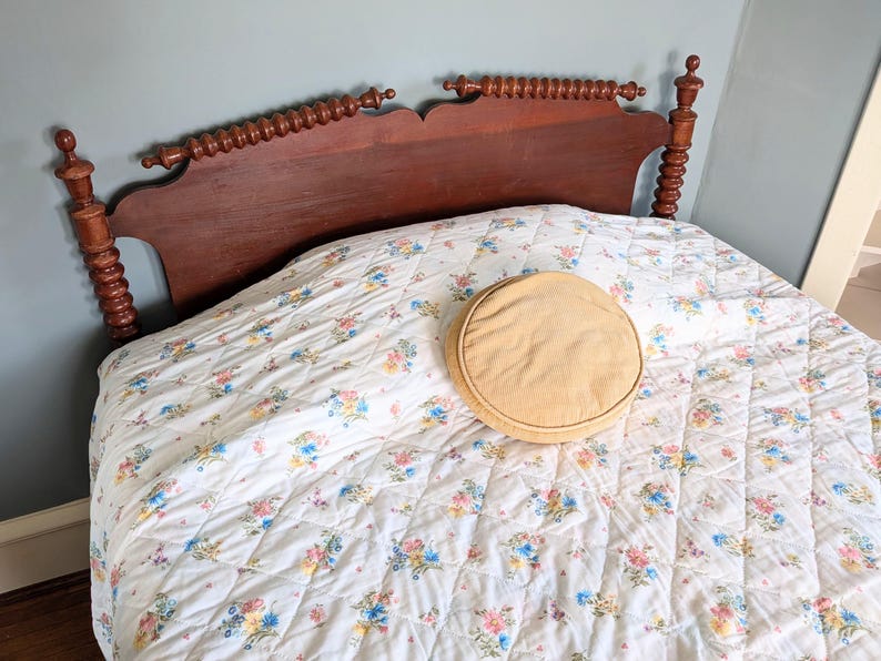 Puede incluir: Un marco de cama de madera con un edred&oacute;n con estampado floral y un coj&iacute;n redondo de color beige. La cabecera es de color marr&oacute;n oscuro con husillos decorativos. El edred&oacute;n presenta un fondo blanco con racimos de flores coloridas.